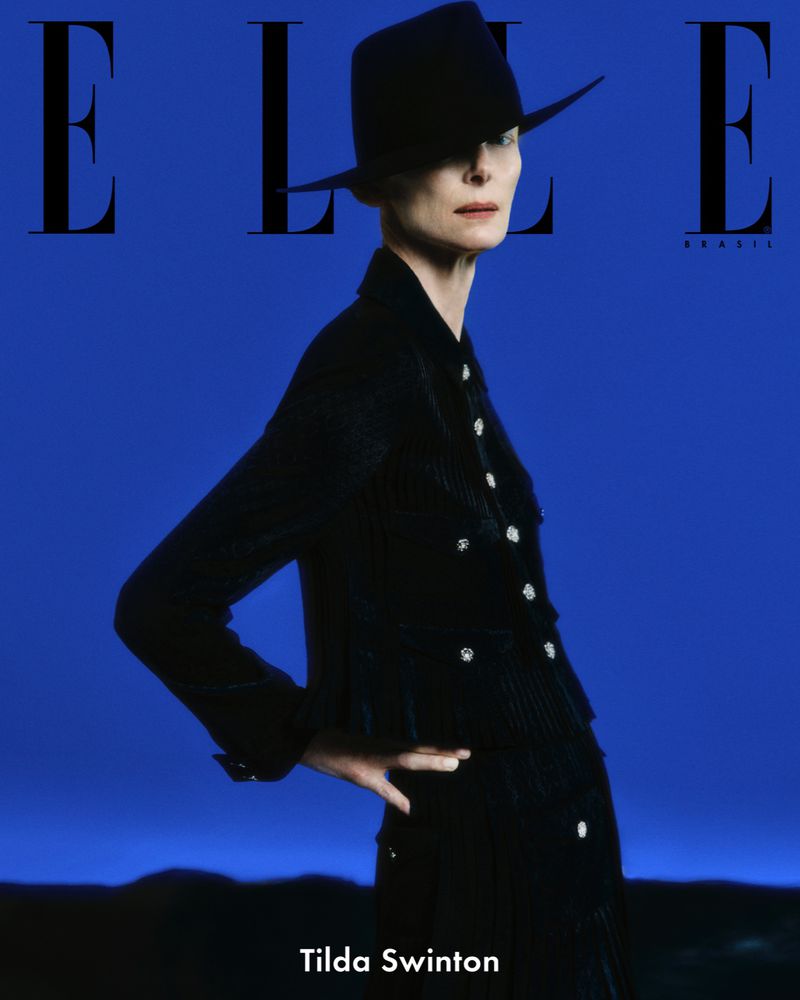 Tilda Swinton in a black suit and hat poses against a blue background on a magazine cover. The 'ELLE Brasil' logo is visible.