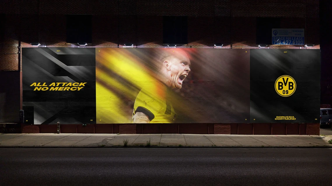 Outdoor billboard at night featuring a soccer player in action alongside sports branding and the slogan 'All Attack, No Mercy'.
