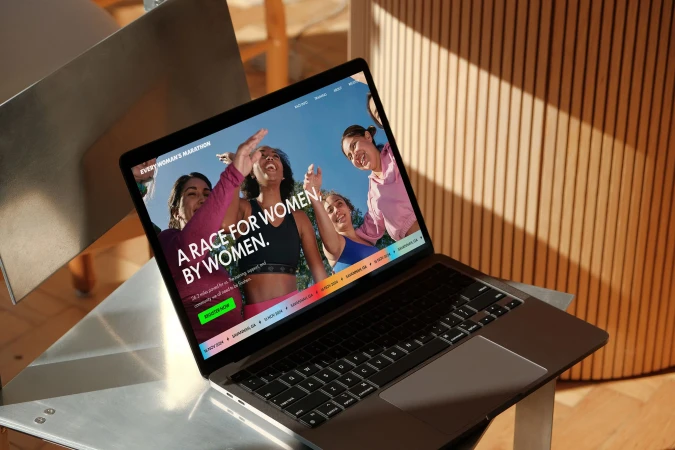 Laptop displays 'Every Woman's Marathon' website featuring diverse women runners, set against wood and metal backdrop.