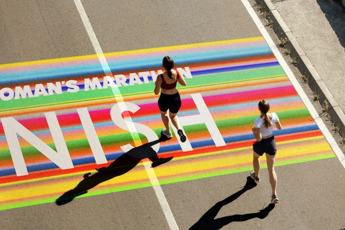 Two women runners approach the finish line on a colorful race course, captured from an overhead shot on a sunny day.