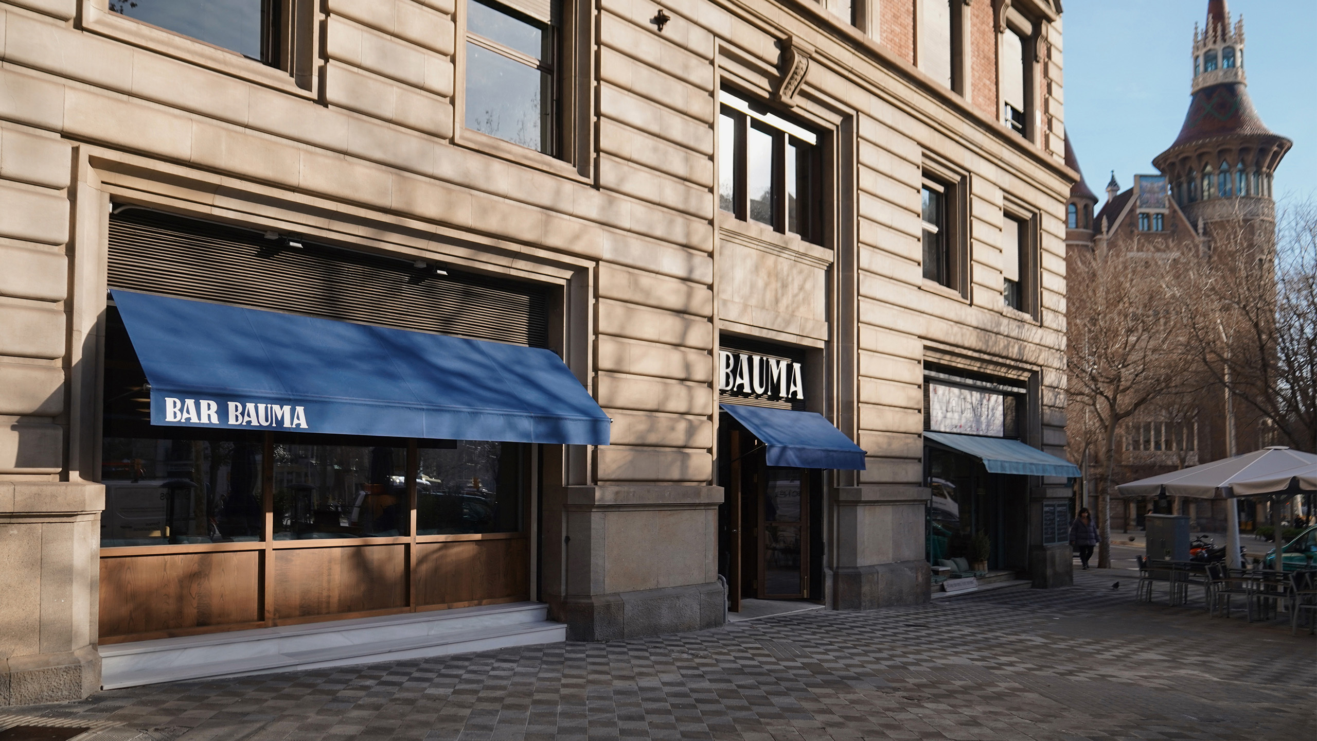 Exterior of Bar Bauma restaurant in an urban environment, featuring modern design with blue awnings and a traditional building facade.