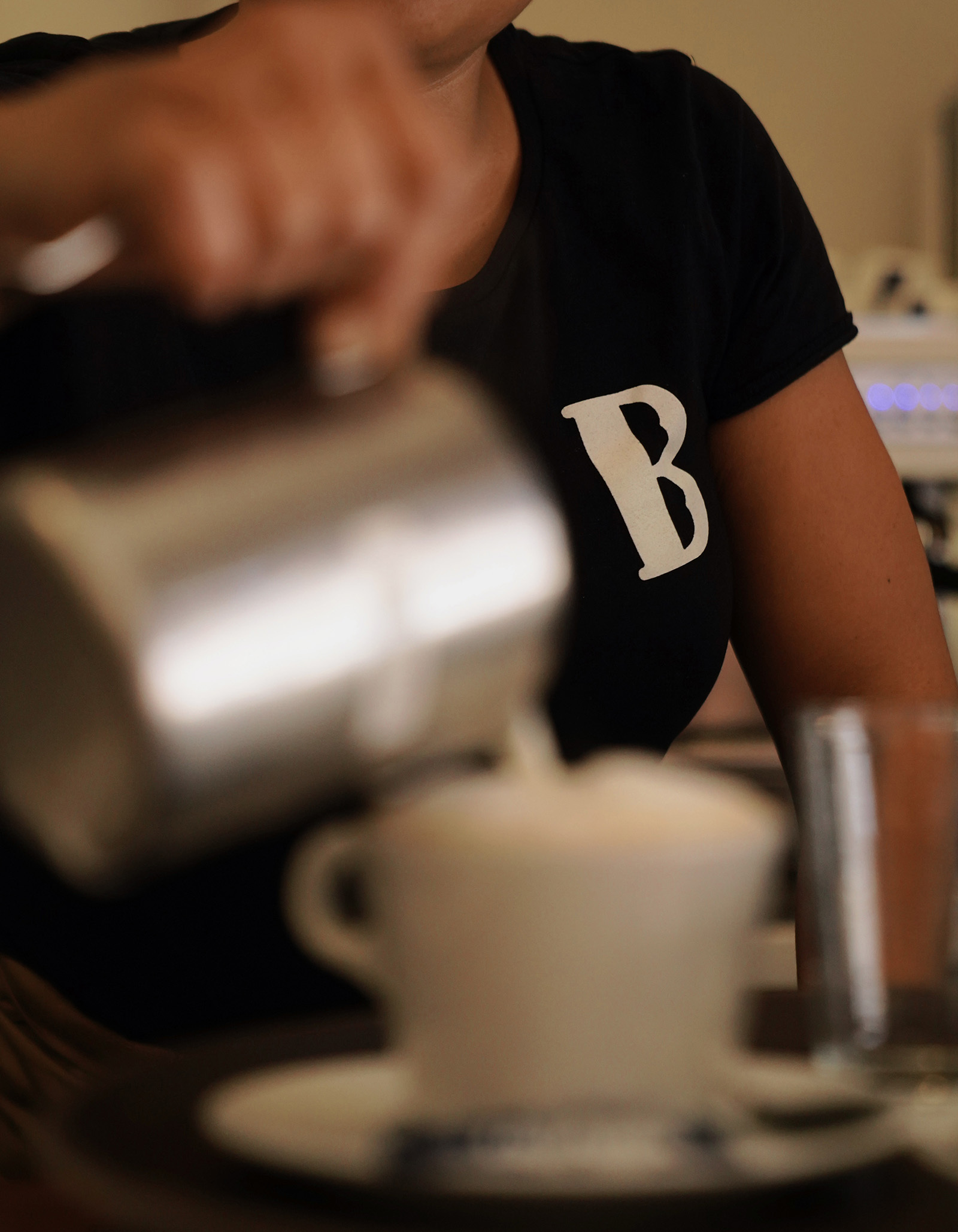 Close-up of barista pouring steamed milk from a stainless steel pitcher into a white ceramic cup with foam on a dark surface.