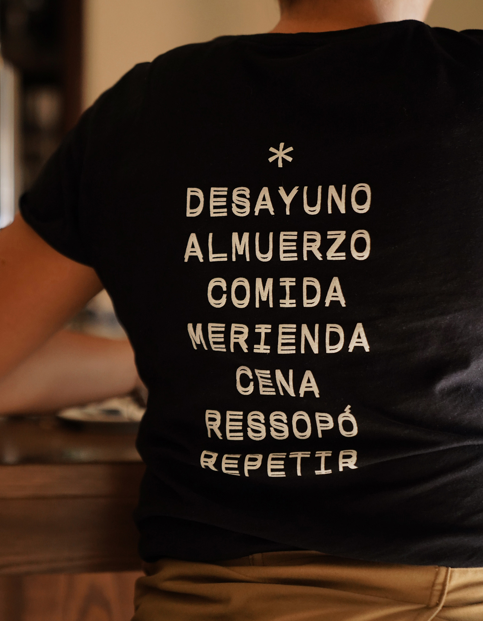 Person wearing a black t-shirt with Spanish text on it stands indoors; the text includes 'Desayuno, Almuerzo, Comida'.