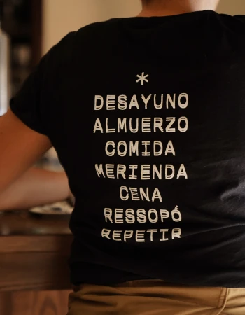 Person wearing a black t-shirt with Spanish text on it stands indoors; the text includes 'Desayuno, Almuerzo, Comida'.