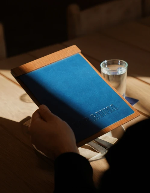 Person sitting at a wooden table, holding a blue and wooden menu, with a glass of water in the background, in warm, natural light.