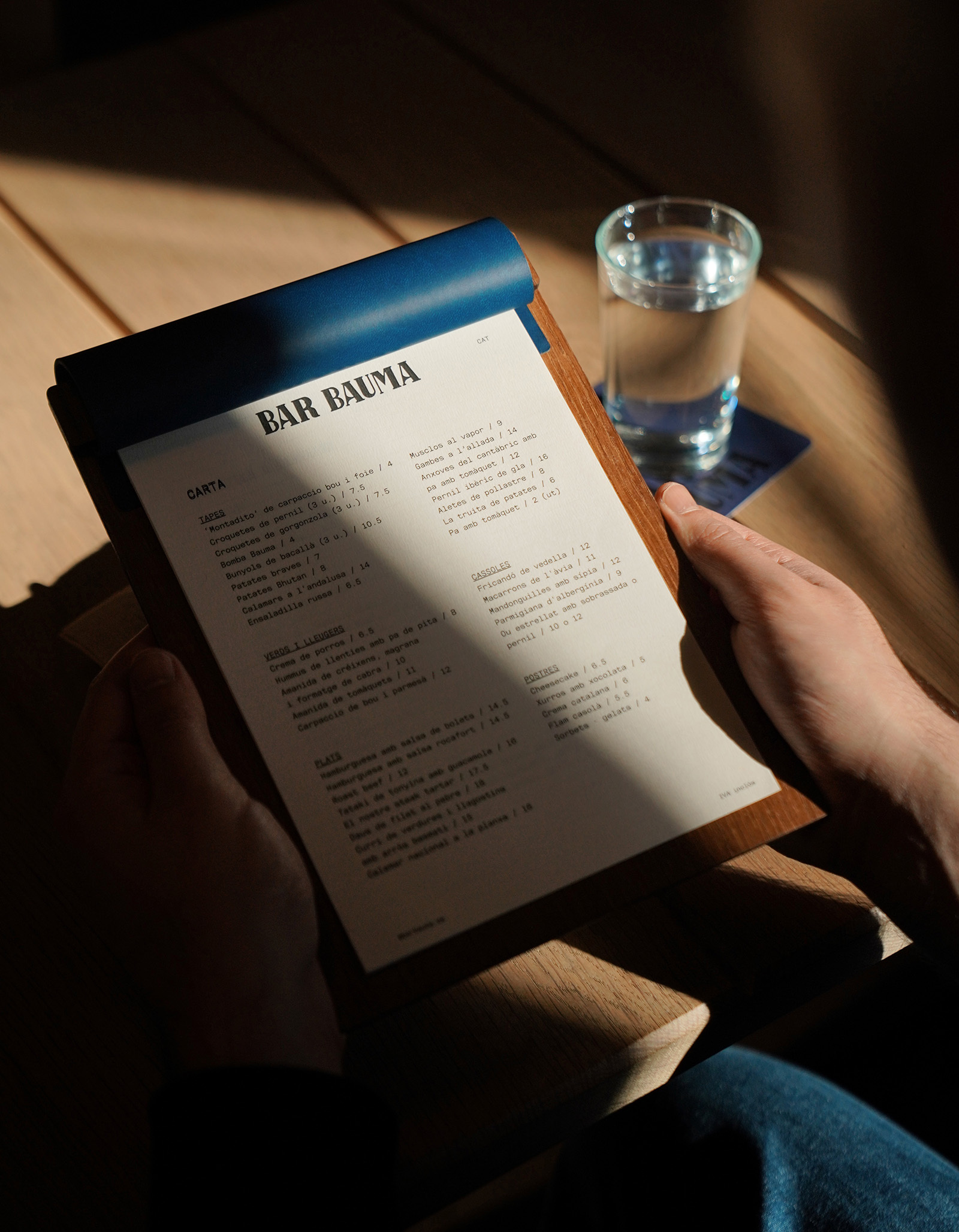 Close-up of a person holding a menu board with a glass of water on the table, sunlight casting shadows. Indoor dining.
