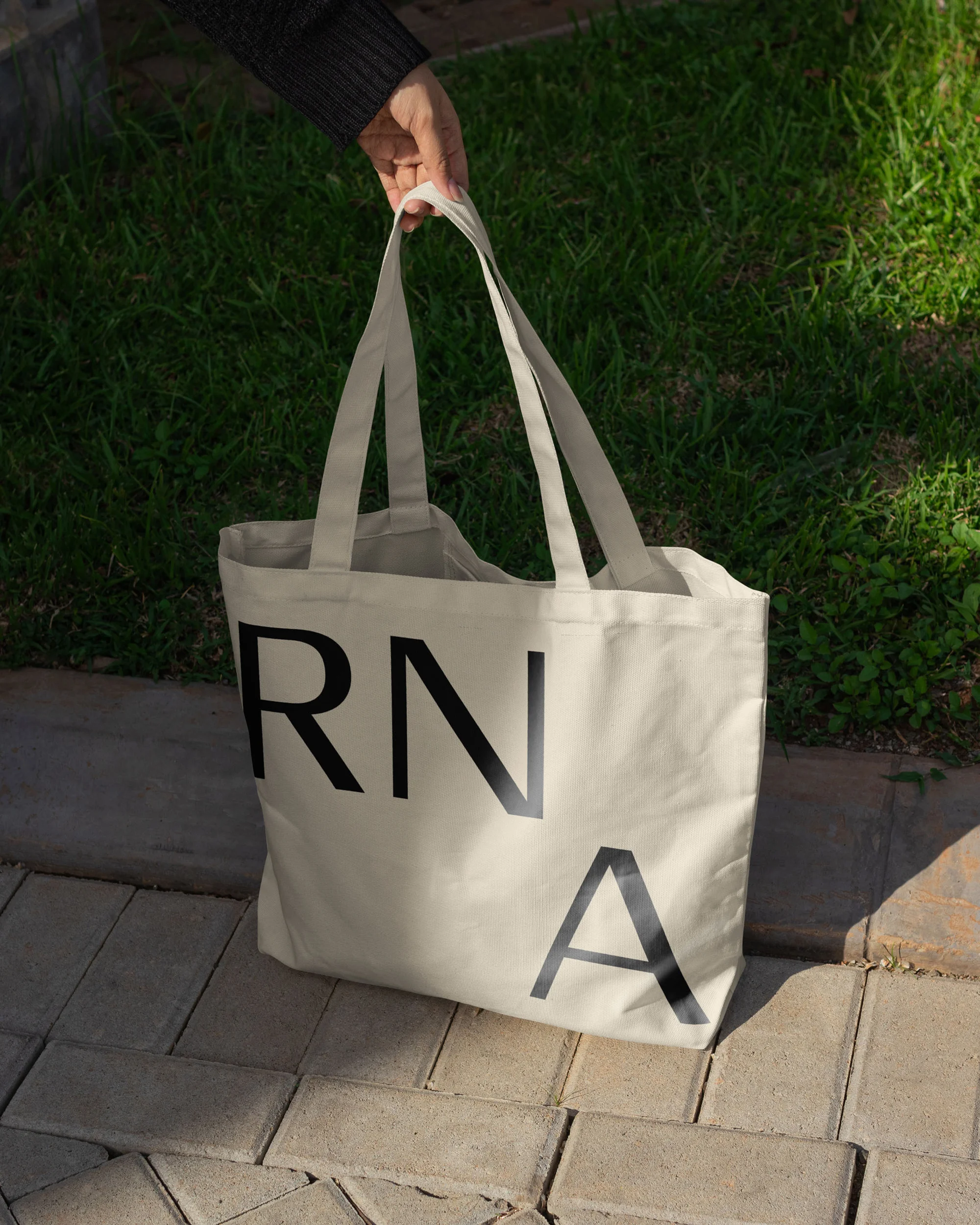 A person holds a white tote bag with the letters RNA printed in black, set against a background of green grass and brick paving.
