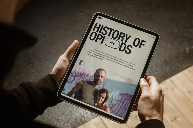 Person holding an iPad displaying an article about the history of opioids with a photograph of a couple with graffiti in the background.