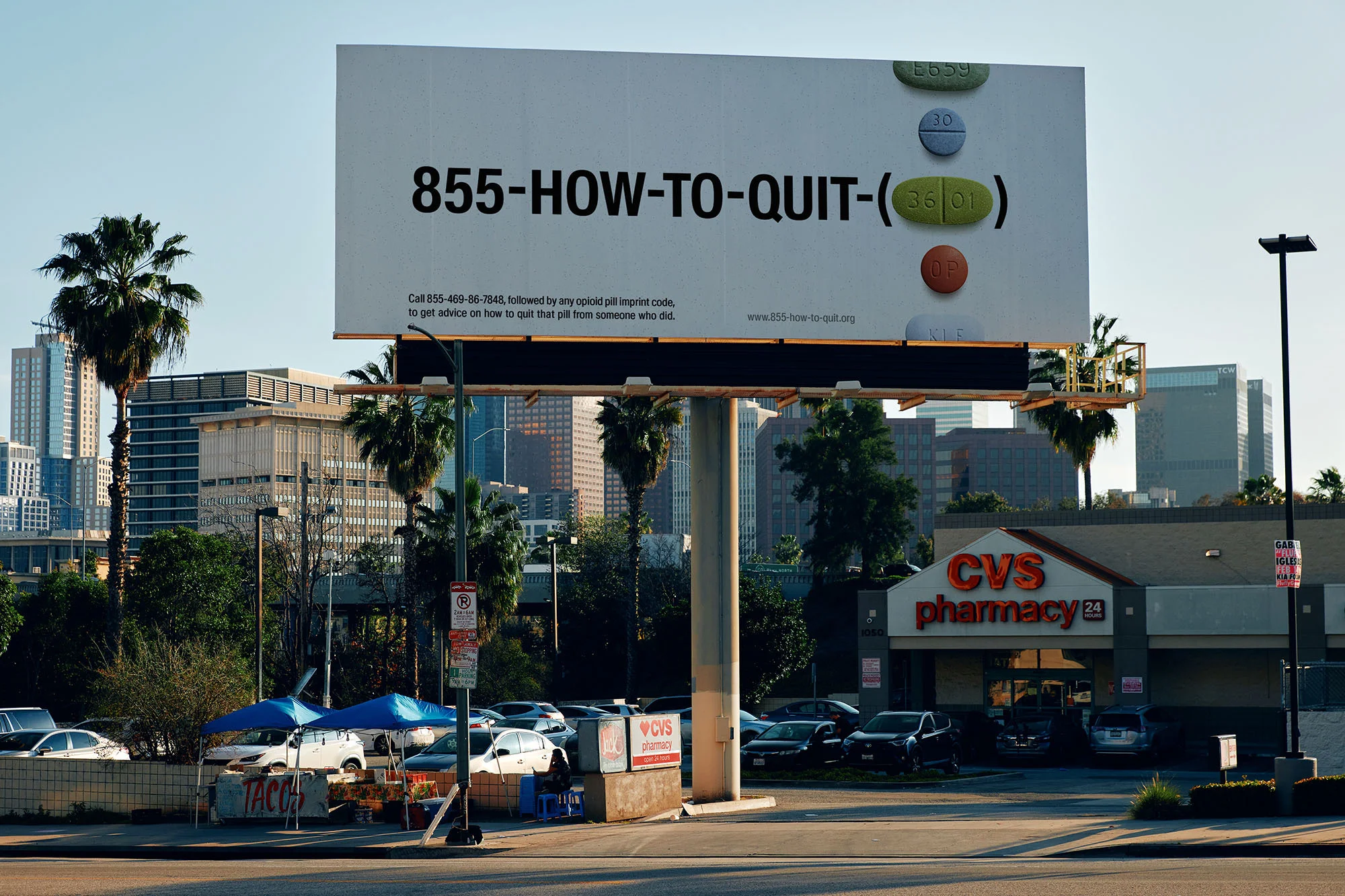 Billboard advertises 855-HOW-TO-QUIT, pharmacy, cars, buildings and palm trees against a clear sky on a sunny day in the city.