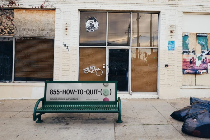 Outdoor view of a vacant white-brick building with boarded-up windows, posters, a green bench, and black garbage bags on the sidewalk.