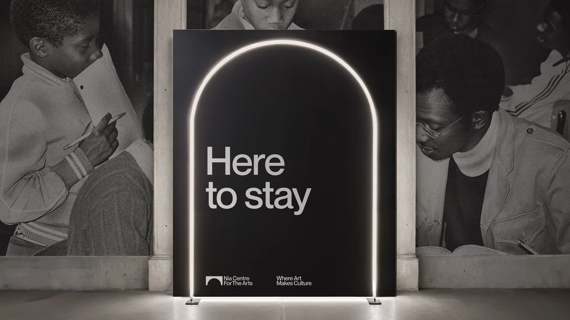 Black advertisement for Nia Centre For The Arts displays 'Here to stay' slogan with arched neon light frame and background photo of students.