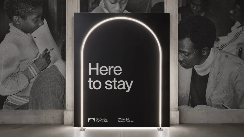 Black advertisement for Nia Centre For The Arts displays 'Here to stay' slogan with arched neon light frame and background photo of students.