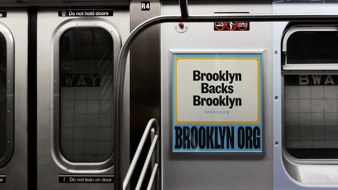 Interior view of a subway car with brushed metal doors and an advertisement for Brooklyn, featuring bold text and a website address.