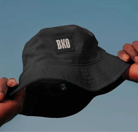 Close-up of a dark-skinned hand holding a black bucket hat with 'BKO' on the front against a clear blue sky background.