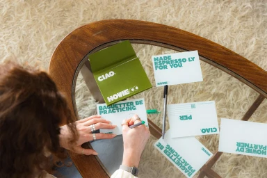 Overhead view of a person writing on a postcard surrounded by branded stationery items on a wooden glass-top coffee table.