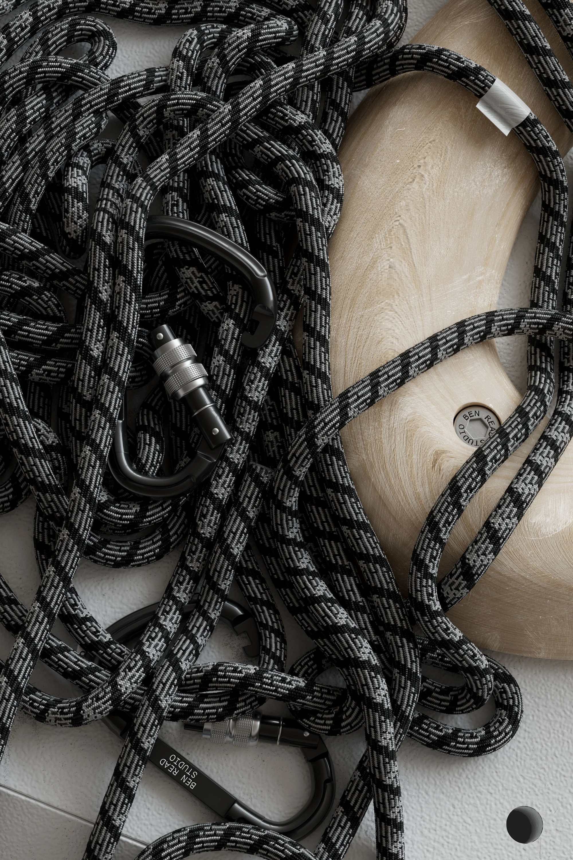 High-angle studio shot of a coiled black-and-gray rope with metal carabiners, alongside a wooden climbing hold, on a white surface.
