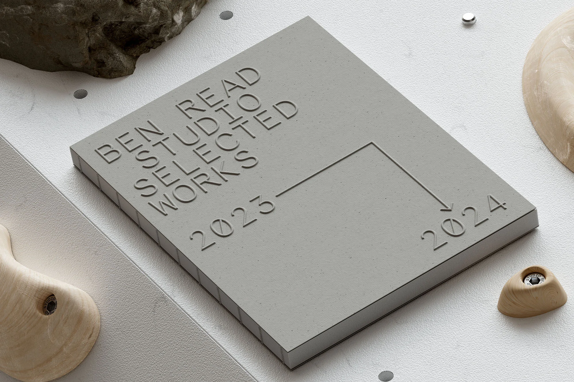 Concrete block with embossed text 'BEN READ STUDIO SELECTED WORKS 2023-2024,' stone, and wooden objects on a white surface.