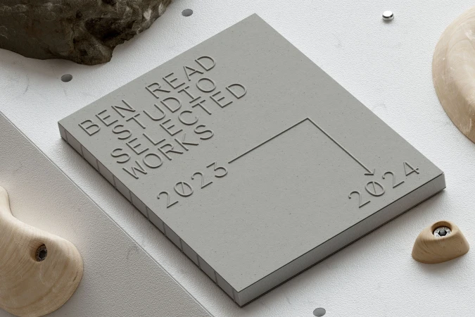 Concrete block with embossed text 'BEN READ STUDIO SELECTED WORKS 2023-2024,' stone, and wooden objects on a white surface.
