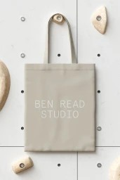 Beige tote bag with the text Ben Read Studio hanging on a climbing wall with textured holds.