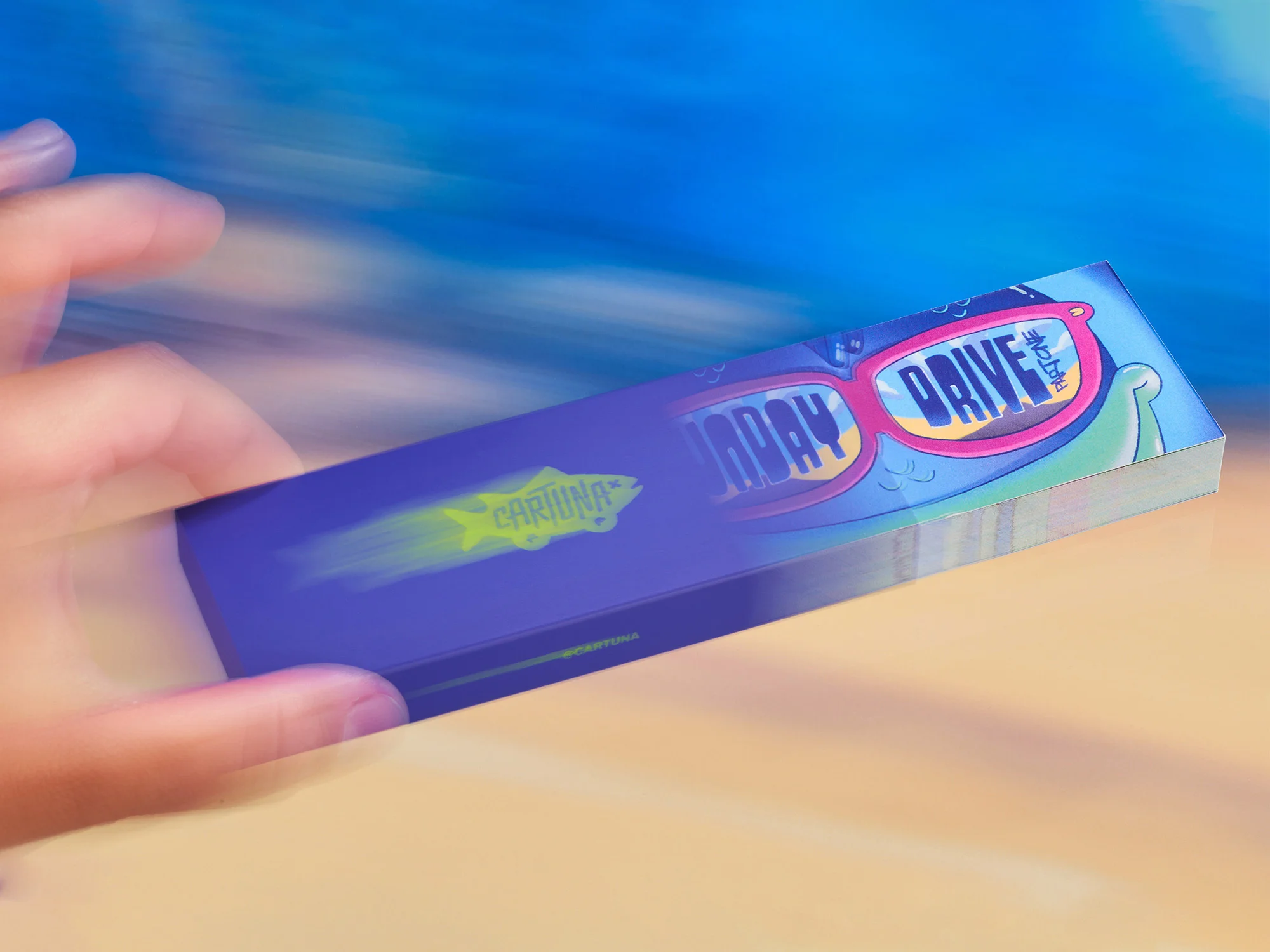 A hand holds a sunglasses case with colorful designs against a blurred background of blue and tan. The case features cartoonish graphics.