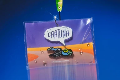 Cartuna branded enamel pin in a clear plastic package hanging from a fishing hook against a vibrant blue background.