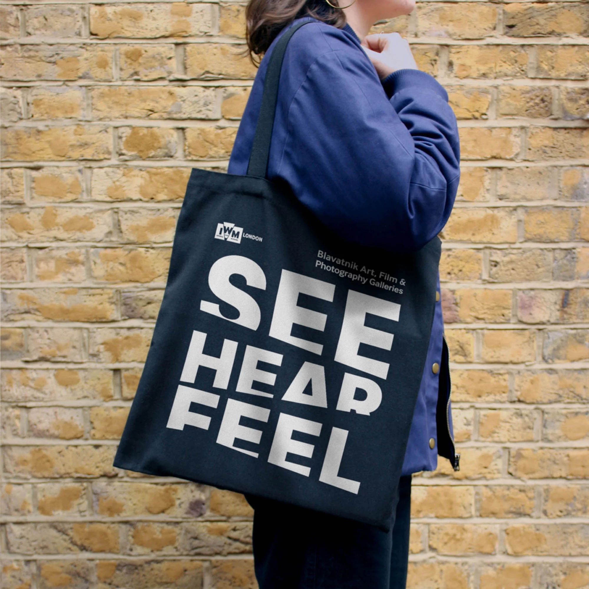 Person carrying a black tote bag with white text 'See Head Feel' against a brick wall. Shoulder-up shot, natural light.