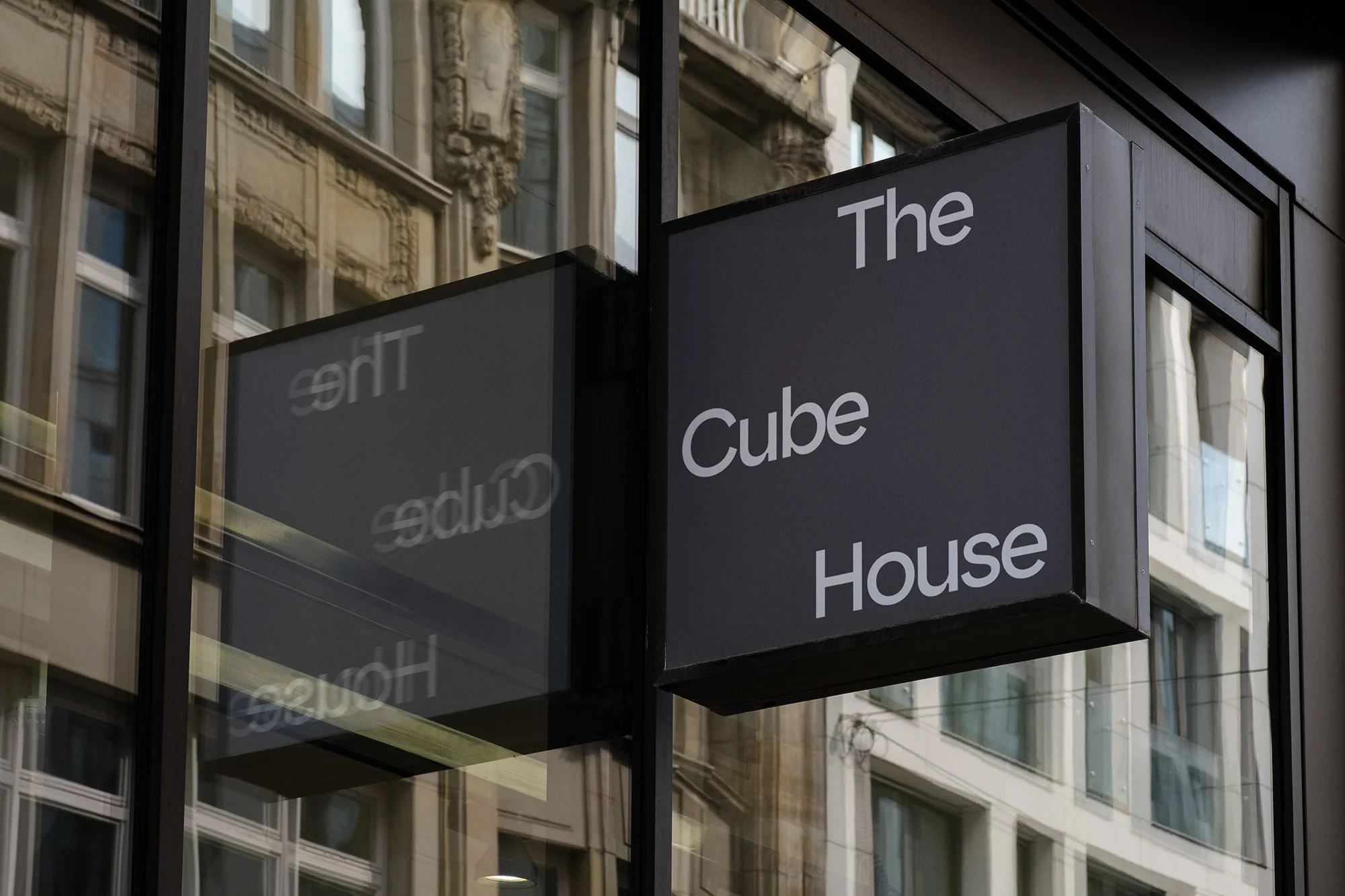 Architectural signage for The Cube House, featuring white text on a dark gray background, reflecting buildings on a city street.