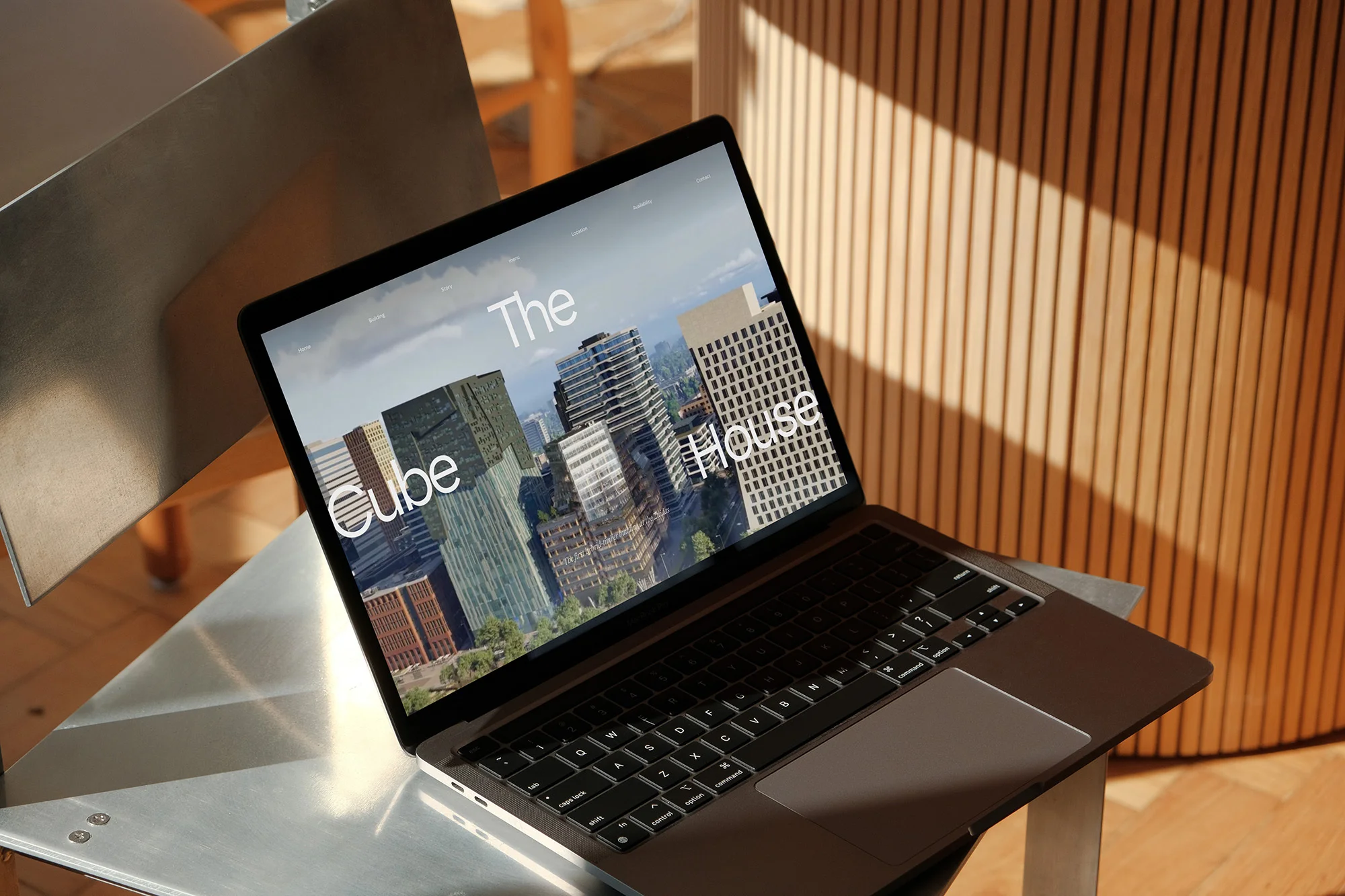 Laptop on a metallic table displaying a cityscape with text "The Cube House" against a vertically-slatted wood wall in daylight.