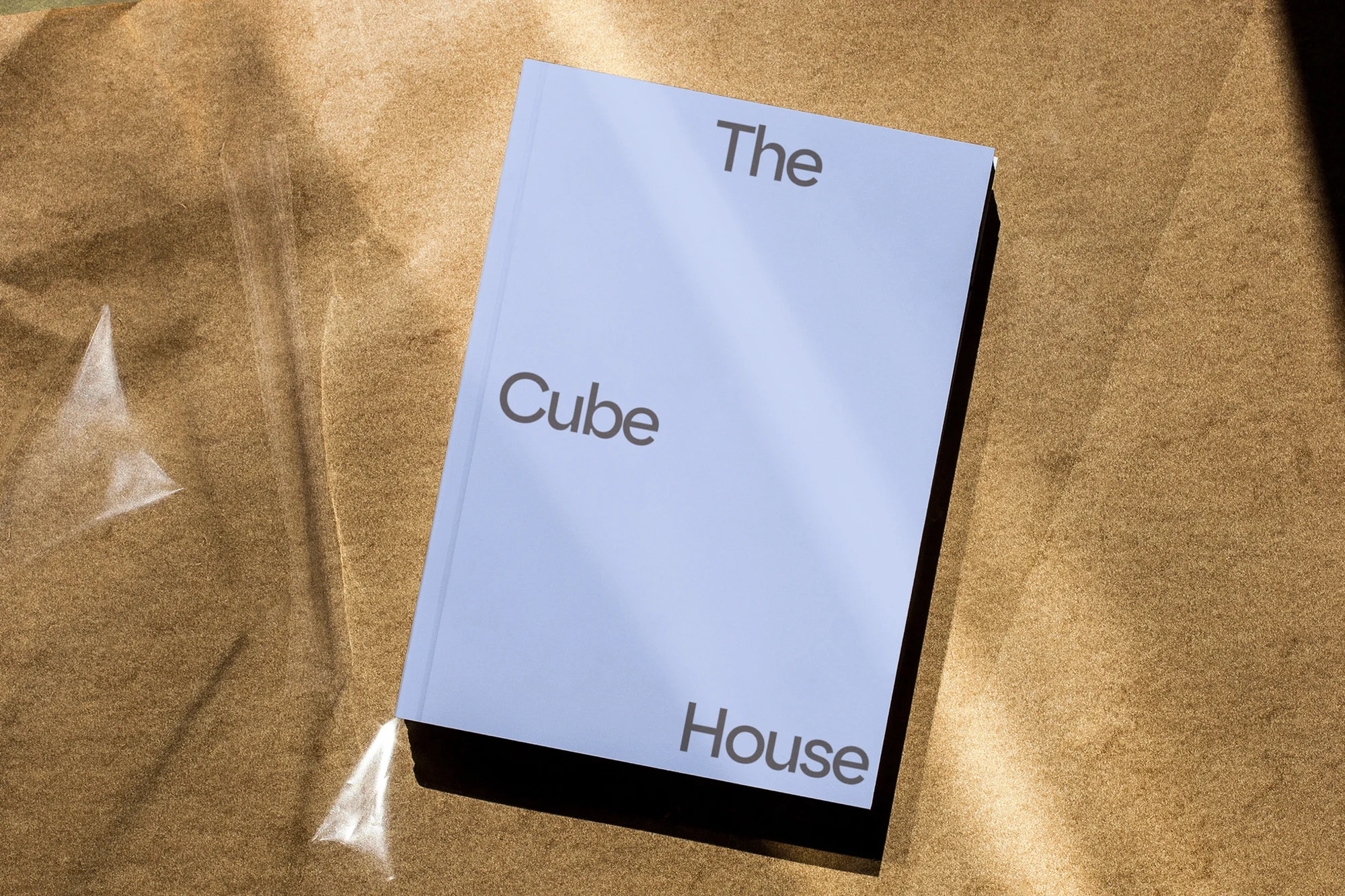 A light blue book titled The Cube House rests diagonally on a crumpled brown paper background under sunlight.