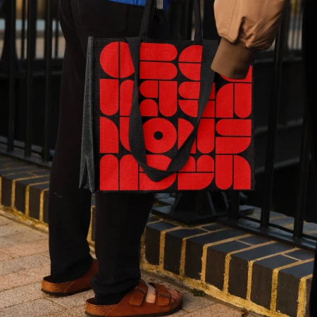 Person carrying a black tote bag with an all-over bold red geometric pattern, paired with black pants and brown sandals, outside.