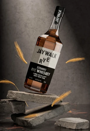Elevated studio product shot of Jaywalk Rye whiskey bottle with rye stalks, set against a textured gray backdrop and gray brick.