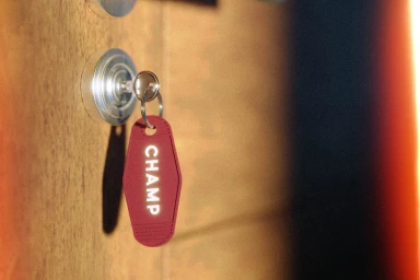 Red motel-style keychain with white CHAMP text hanging in a keyhole on a wooden door, photographed with warm lighting and film grain.