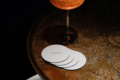 Round white coasters with CHAMP branding resting on a textured bronze tray beside a cocktail glass