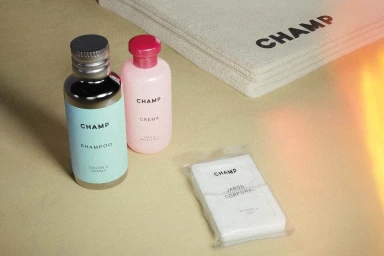 CHAMP branded cosmetic bottles, soap, and folded towel on a textured neutral surface with warm light flare