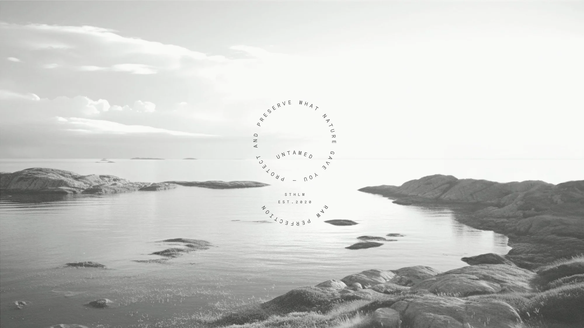 Monochrome landscape shot of rocky shoreline, calm water, and cloudy sky. A circular text overlay with brand name and tagline.