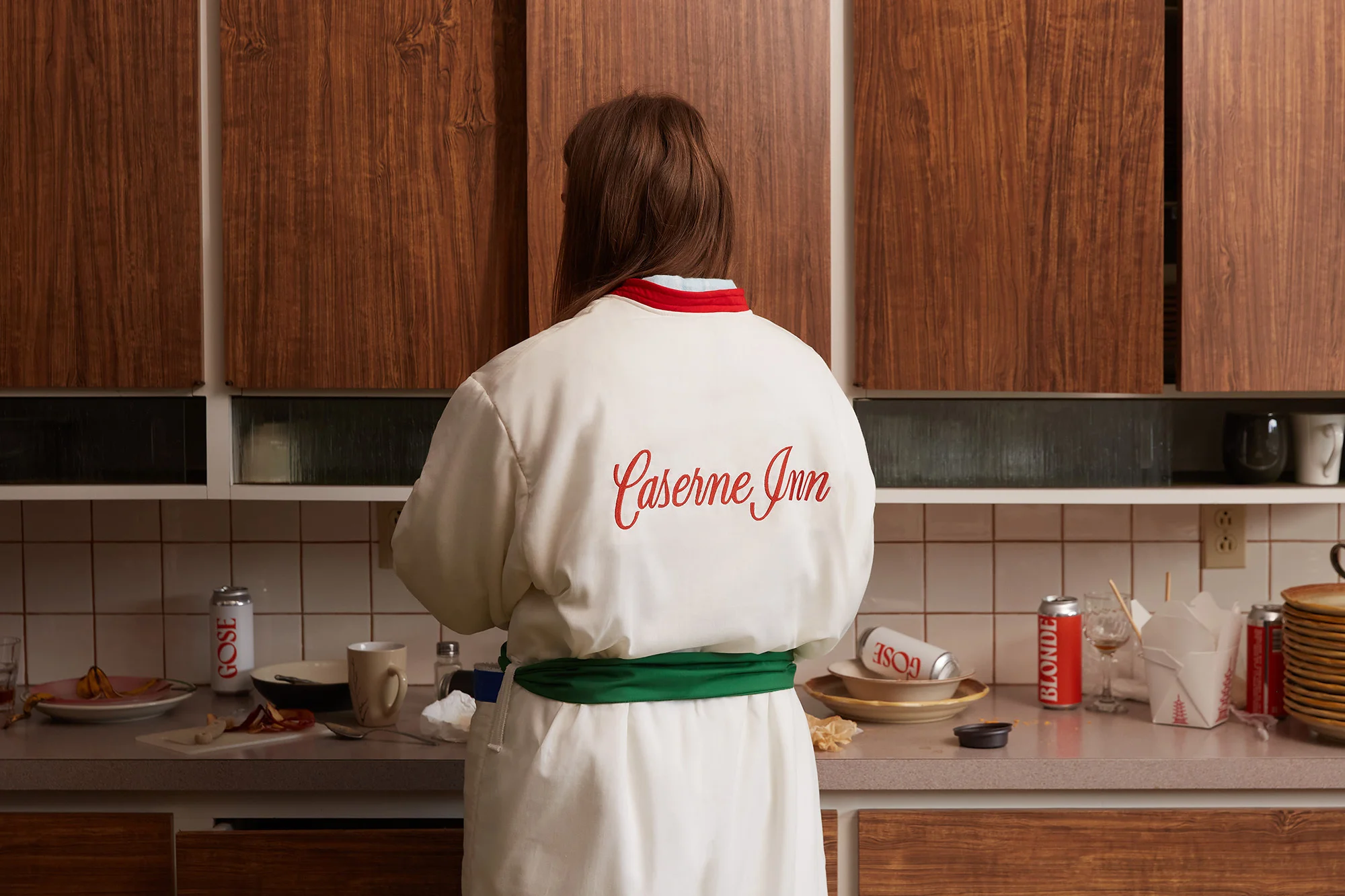 Back view of a woman wearing a white robe with 'Caserne Inn' embroidered, standing in a kitchen with dirty dishes and drink cans.