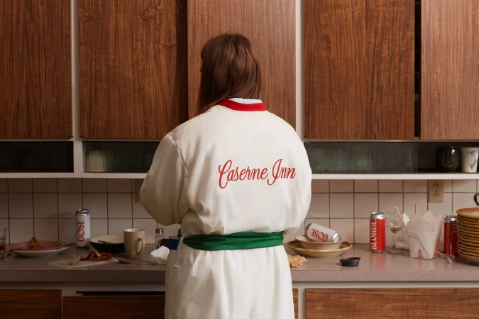 Back view of a woman wearing a white robe with 'Caserne Inn' embroidered, standing in a kitchen with dirty dishes and drink cans.