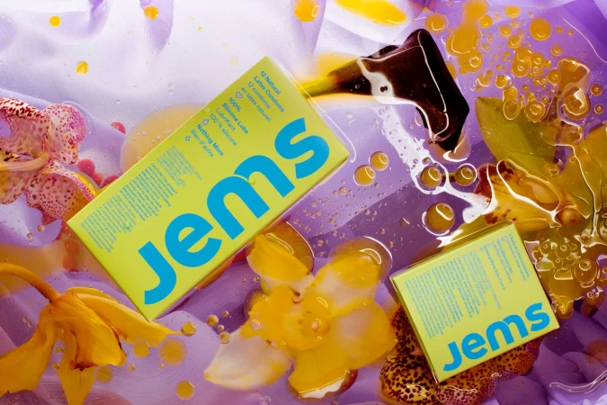Two Jems condom packages are arranged with orchid flowers, droplets, and yellow oil against a light purple background in a studio setting.
