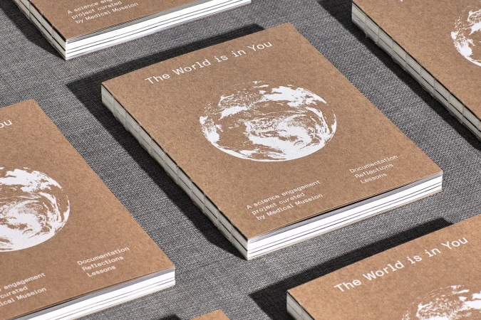 Close-up of stacked journals with the text 'The World is in You' and a graphic of Earth on a tan paper cover against a gray backdrop.