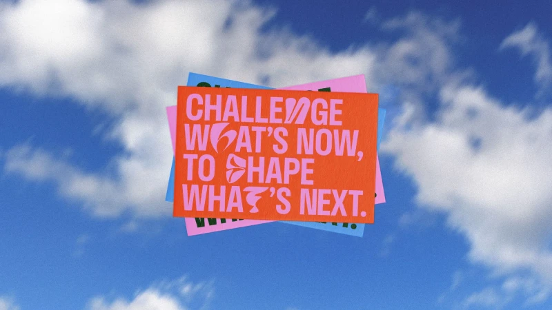 Text on colorful cards against a bright blue sky with clouds displays the message 'Challenge What's Now, to Shape What's Next'.