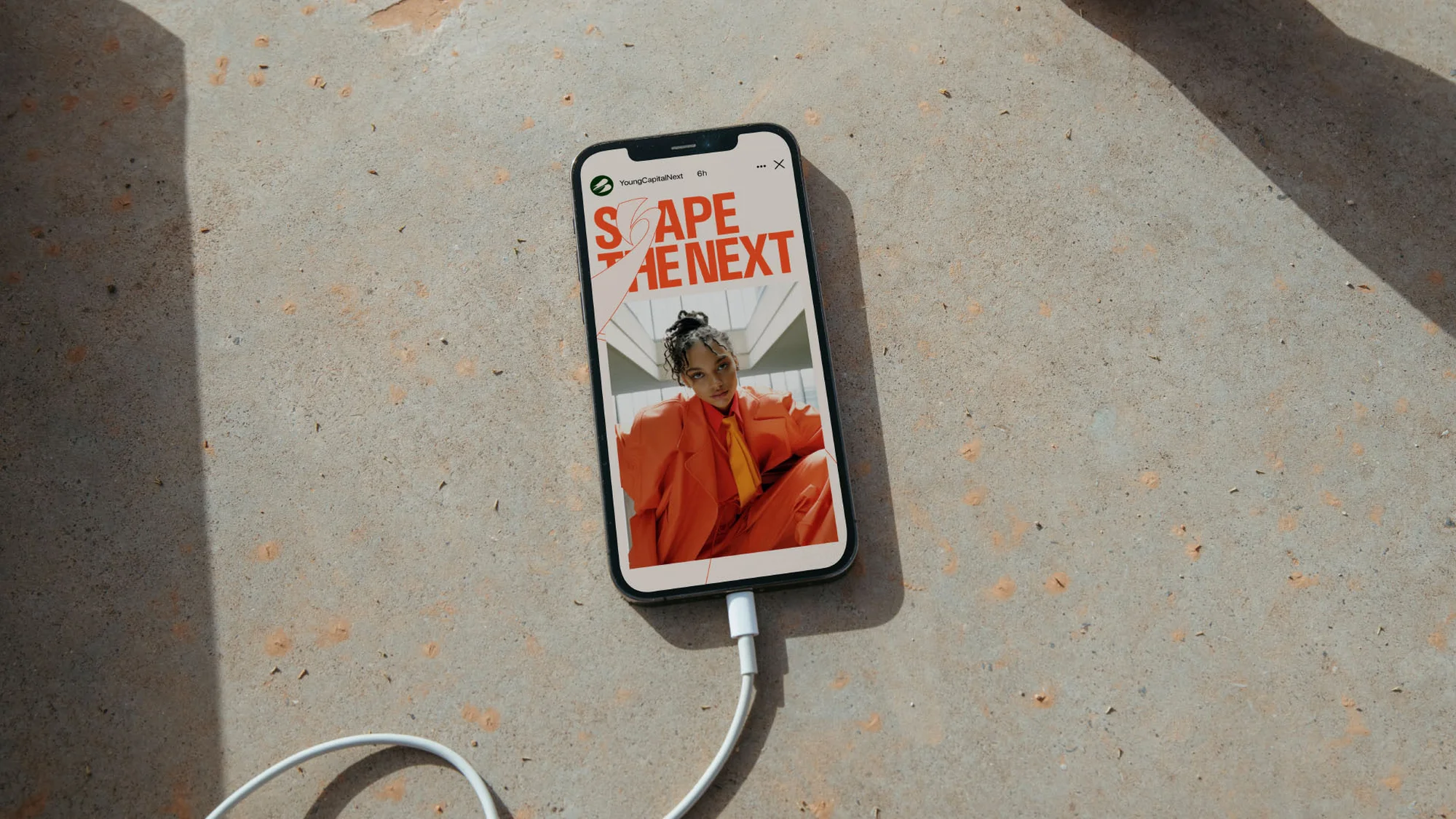 Smartphone displays a striking woman in an orange suit against a gray concrete surface, connected to a white charging cable.