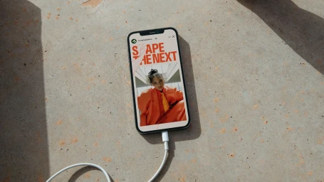 Smartphone displaying a YoungCapital NEXT brand story resting on a textured concrete surface with a white charging cable attached.