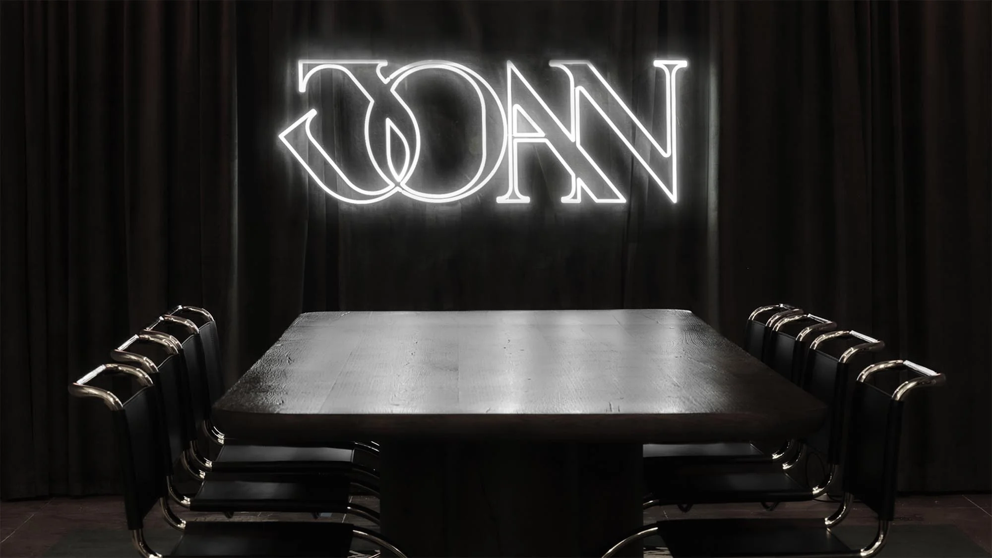 Illuminated neon sign that reads 'JOAN' centered above dark wood conference table surrounded by black and chrome chairs against black curtain backdrop.