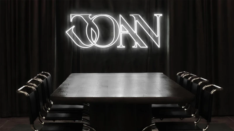 Illuminated neon sign that reads 'JOAN' centered above dark wood conference table surrounded by black and chrome chairs against black curtain backdrop.