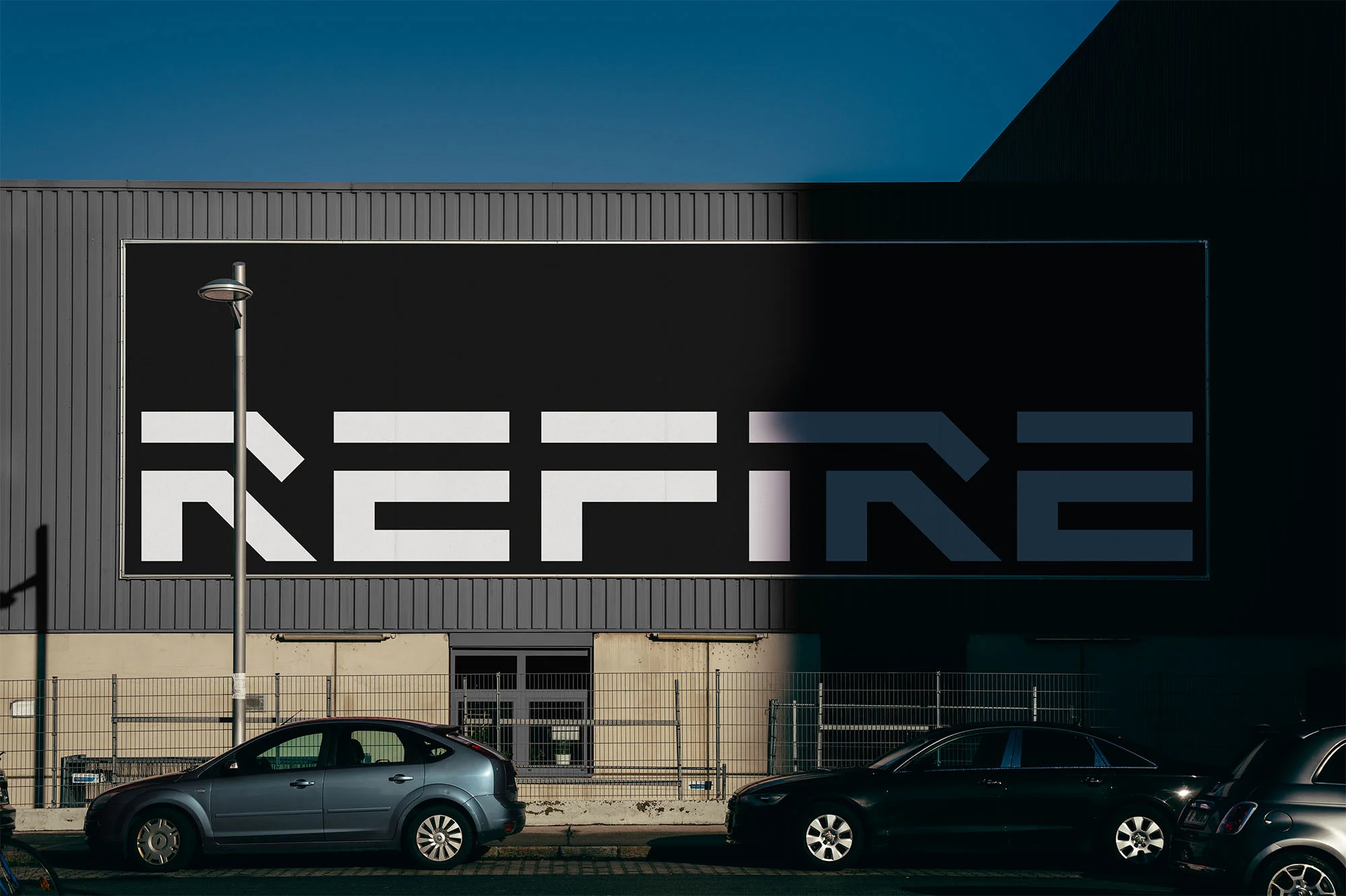 Exterior of a grey building with a large REFIRE sign in white and black, parked cars, street lamp, clear blue sky, and shadows.
