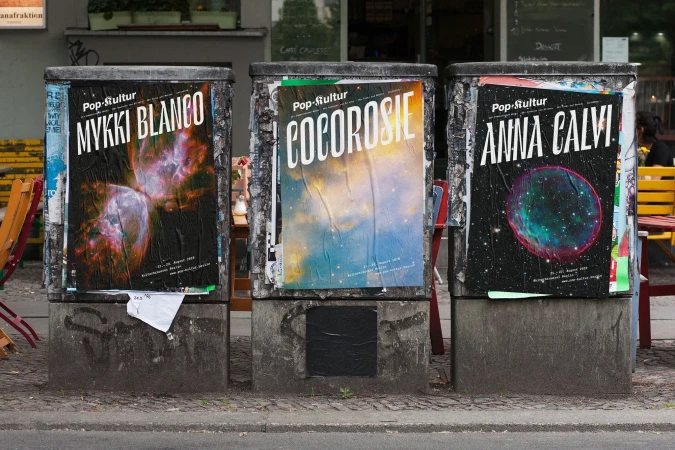 Three music posters on concrete pillars in an urban setting, featuring MYKKI BLANCO, COCOROSIE, and ANNA CALVI.