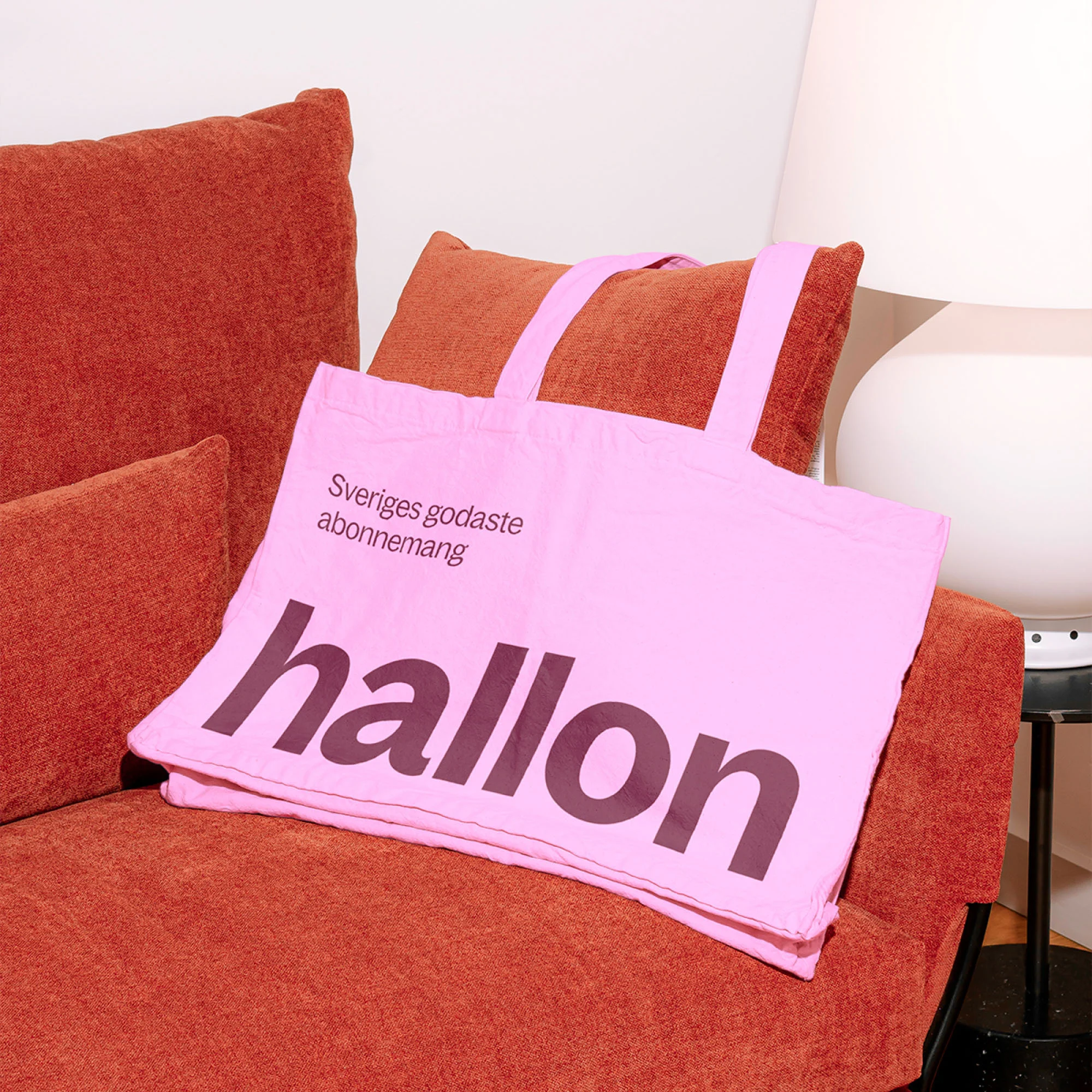 Pink tote bag featuring the word 'hallon' displayed on an orange armchair beside a white lamp and small black table.