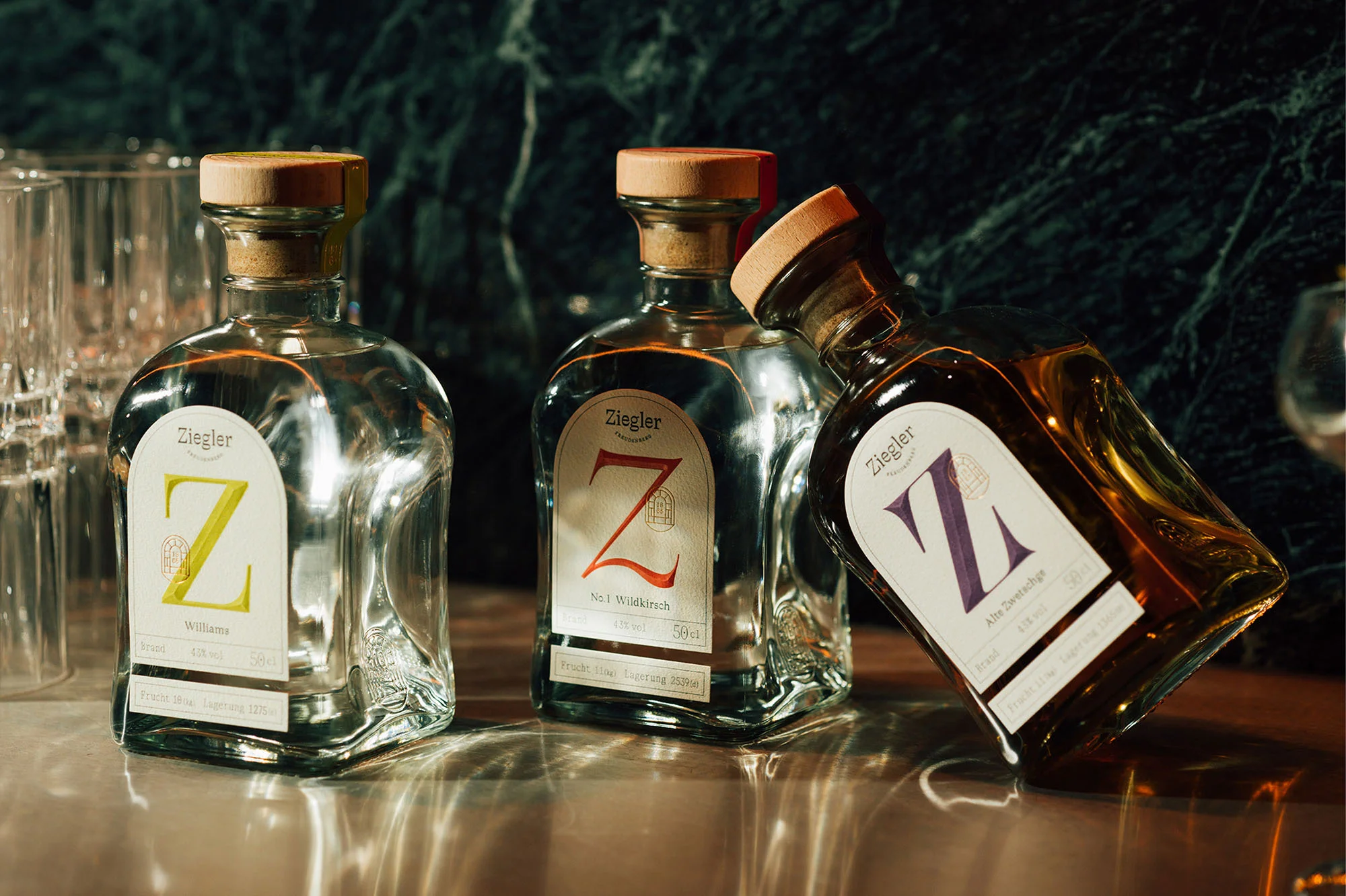 Three Ziegler brand spirit bottles with cork stoppers on a reflective surface, against a dark marble background. Food & beverage shot.