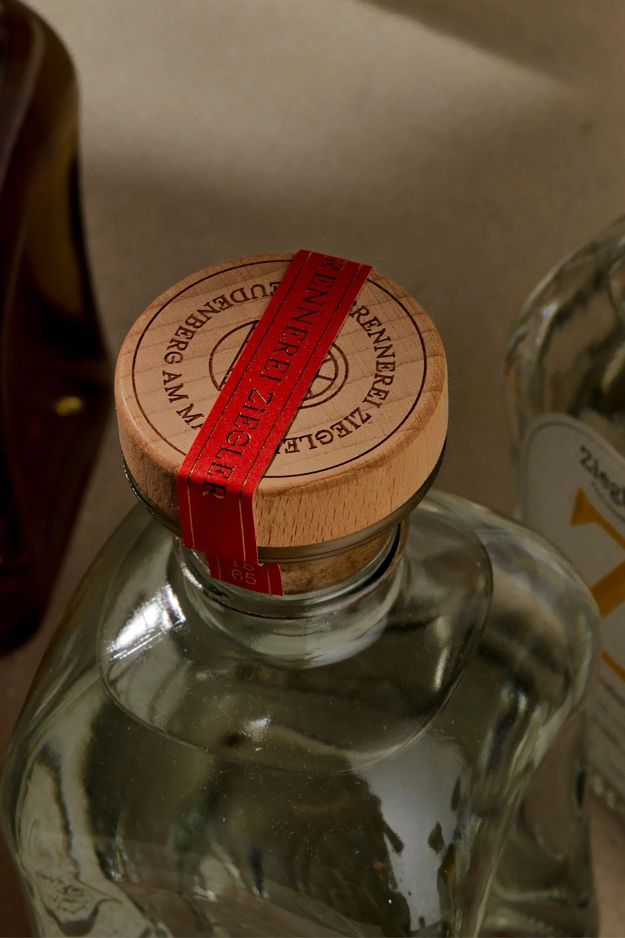 Close-up of a clear glass bottle with a wooden cork stopper, sealed with a red band, showcasing branding details on the lid.