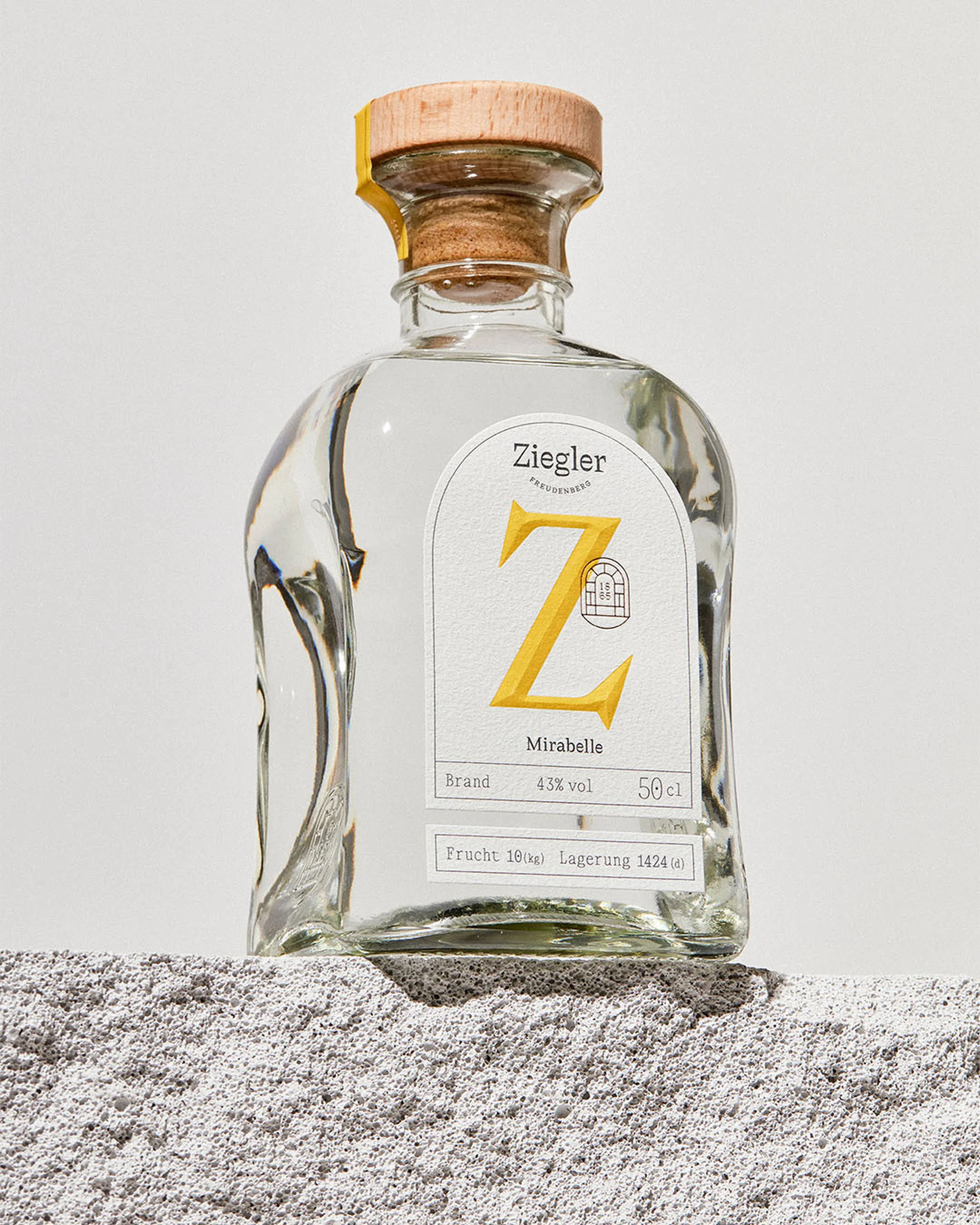 Elegant clear glass bottle of Ziegler Mirabelle brandy with wooden stopper on a textured gray surface against a white background.